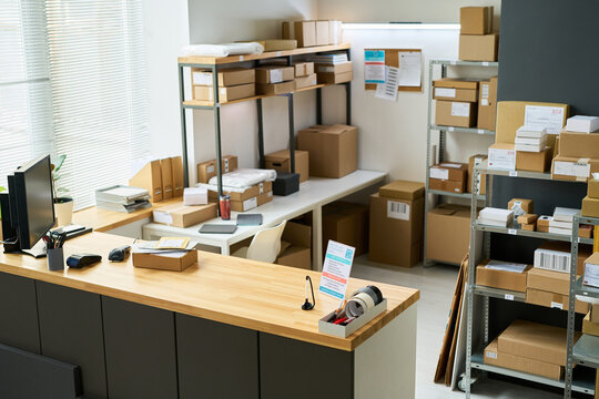 Empty modern shipping office showing organized shelves filled with cardboard boxes, packing materials, computer monitor on counter, barcode scanner, tape dispensers, daylight streaming through window