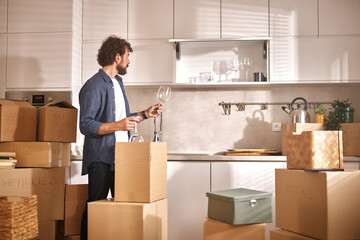 A man stands in a newly empty kitchen, unpacking glassware from boxes. Sunlight streams through the...