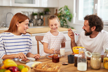 A woman and man share a meal with a young girl, all seated at a wooden table. They are smiling and enjoying a variety of breakfast foods, creating a warm atmosphere.