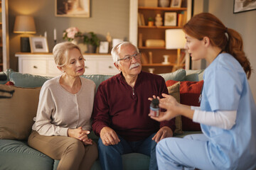 A female nurse wearing scrubs holds a medication bottle and speaks to an elderly couple seated on a couch in their home. She is providing home health care and explaining medication instructions.