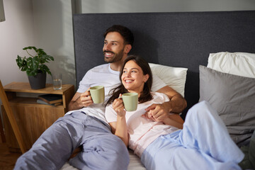 A couple shares a joyful moment in bed, holding warm cups and smiling at each other in a stylish, minimalist bedroom. Soft lighting enhances the relaxed atmosphere.