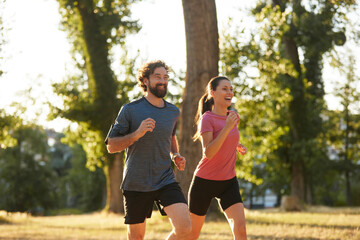 Fototapeta premium Two runners smile and practice their fitness routine in a sunlit park, enjoying their morning exercise amidst lush greenery and warm weather.