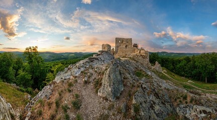 Ruins of an old stone castle on a rocky hilltop surrounded by forest and mountains, lit by the warm light of a colorful sunset in summer.