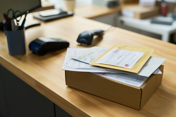 Stack of parcels and envelopes resting on wooden office desk, including cardboard box and various documents, background showing payment terminal and office supplies in workspace