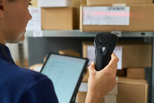 Caucasian young adult woman scanning barcode on cardboard box with handheld scanner while holding digital tablet in warehouse storage area, focusing on inventory management process