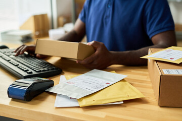 Black man working at counter handling cardboard box and envelopes, using computer keyboard and payment terminal, processing packages for shipping or delivery service in office setting