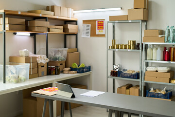 Shelves filled with cardboard boxes, canned goods, bottles, and plastic containers standing in organized storage room, digital tablet and paperwork lying on table in foreground