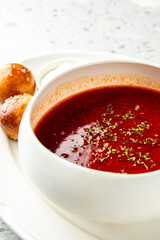 Tomato soup with herbs served in a white bowl on a plate, accompanied by glazed bread rolls and sour cream. Minimalist food presentation on a speckled countertop.
