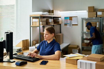 Caucasian young adult woman working at computer in warehouse office, typing on keyboard while surrounded by packages, Black man in background sorting parcels on shelves, modern workspace