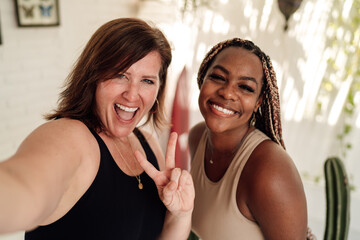 Happy diverse women laugh and make peace sign while taking selfie, reflecting joy, tech interaction and vibrant connection through mobile culture.