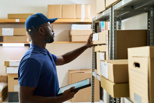 Black man young adult organizing cardboard boxes on storage shelf while holding digital tablet in hand, working in warehouse or stockroom, focusing on inventory management tasks