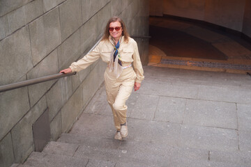 Smiling woman walking up stairs while holding handrail. City life in comfortable beige jumpsuit and colorful scarf. Middle-aged woman (50 years old)