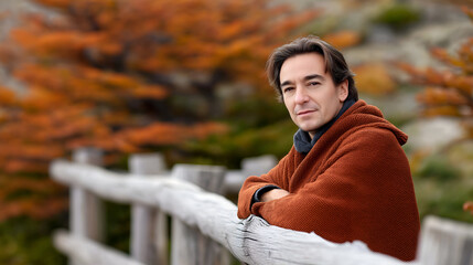 Man wrapped in a cozy orange blanket leans against a wooden fence, surrounded by vibrant autumn foliage, capturing a serene moment in nature's beauty and tranquility