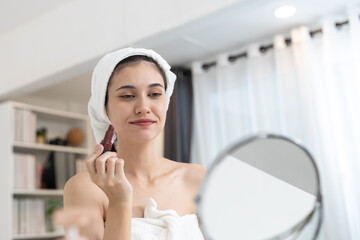 Young women wrapped in towel applying lipstick while smiling at mirror in bright room. Concept of...