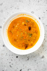 Homemade vegetable soup in white bowl on gray countertop. Rich orange broth with carrots, potatoes, beans, herbs. Healthy, rustic, freshly made meal. Top view, natural light