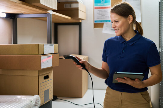 Caucasian young adult woman scanning cardboard boxes with barcode scanner while holding digital tablet in warehouse storage room, smiling and focusing on inventory management process