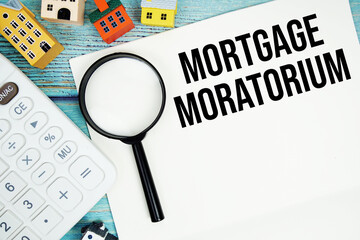 Notepad with "Mortgage Moratorium" with Calculator, magnifying glass and house model on wooden background