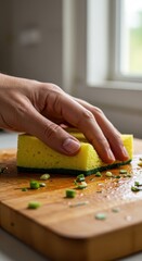Fototapeta premium A hand cleans a wooden cutting board with a yellow sponge and chopped green onions.