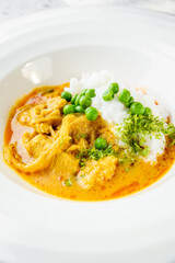 Plate of creamy chicken curry with white rice and green peas, garnished with fresh herbs. Vibrant colors and appetizing presentation on a white dish.