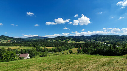 Wide countryside panorama with green fields, forests, and scattered houses stretching toward distant hills and fluffy clouds.