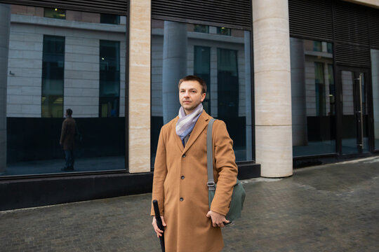 Man in a brown coat stands confidently outside modern building reflecting cityscape. A 37 years old Caucasian man goes to work. He is an office worker, IT specialist, executive, or manager.