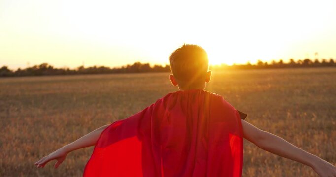 Superhero child silhouette in red cape runs toward sunrise outdoors with arms outstretched as if flying. child freedom, imagination, motivation, and aspiration in open landscape with dramatic sunlight