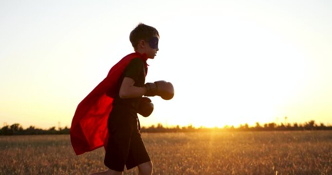 motivation child superhero in red cape and boxing gloves runs through meadow golden hour outdoors. superhero child shows perseverance, ambition, and strong will. Cape flies in wind, inner drive.