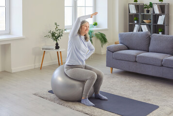 Old senior woman actively exercising indoors at home, sitting on large fitness ball, stretching,...