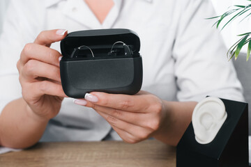 Doctors hands holding an open charging case with two digital hearing aids. Modern assistive technology for hearing health and daily use