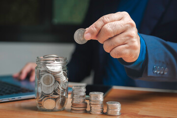 Businessman hand putting coin into glass jar with stacked coins on desk, concept of saving money, financial growth, investment, budgeting, wealth management, and personal finance planning.