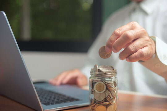 Close up of businessman hand putting coin into glass jar with laptop on desk, concept of saving money, financial planning, investment, economy, wealth management, and personal finance growth.