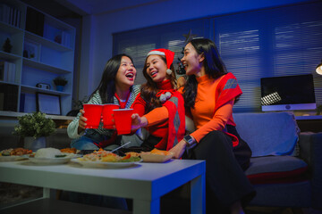 Young Asian women friends celebrating Christmas at home, toasting with red cups, laughing and...