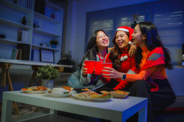 Young Asian women friends celebrating holiday season, toasting with cups at a Christmas house party, enjoying food and friendship