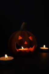 Carved pumpkin glows in the dark: a jack-o-lantern with triangular eyes and toothy grin, lit by candles on a black background; moody studio still life for autumn holiday themes.