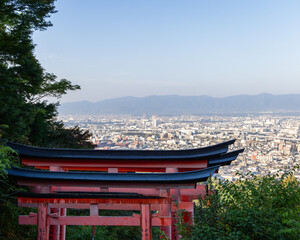 View from the sacred forest of Mount Inari in Kyoto, Japan, showing vermilion torii roofs in the foreground and the urban landscape stretching into soft morning haze