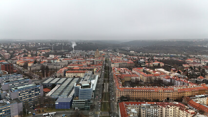 Aerial winter view of dense historic blocks with red-tiled roofs and a curving perimeter, broad avenues streaming traffic, distant parkland and a rail corridor fading into soft overcast haze. © VladyslavShcherbakov