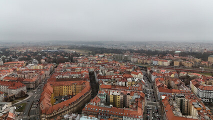Aerial winter view of dense historic blocks with red-tiled roofs and a curving perimeter, broad avenues streaming traffic, distant parkland and a rail corridor fading into soft overcast haze. © VladyslavShcherbakov