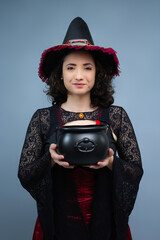Friendly mid-adult witch holding a black cauldron with both hands looking at camera. Ideal for Halloween trick or treat, witches brew, potion, and fairy tale character. Studio portrait with copy space
