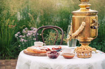 A golden samovar with bagels, tea cups, homemade pie, and jam on a white tablecloth set for tea in a garden.