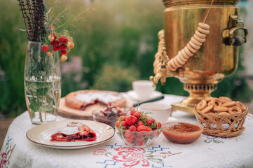 A rustic tea setting with golden samovar, fresh strawberries, jam, pie, and bagels arranged on an embroidered tablecloth outdoors.