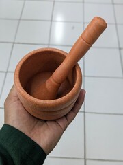 A hand holds a small, light brown, clay mortar and pestle, ready for grinding spices or herbs.