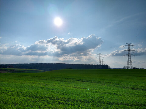 Bright afternoon over rolling green farmland with dramatic clouds and tall transmission towers on the horizon.