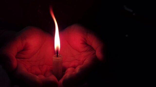 Close-up of a hand holding a lit candle in the dark background.