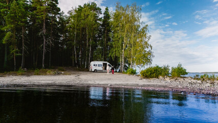 A tranquil lakeside scene at Lake Saimaa featuring a campervan, lush trees, and calm waters during a beautiful day. Perfect for outdoor adventure and relaxation in nature of Finland