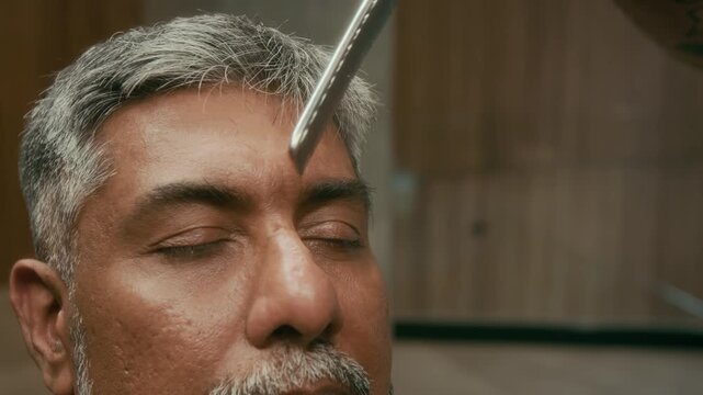 Close-up of hands of tattooed young professional barber combing and trimming with scissors eyebrows for mature Indian man, sitting with closed eyes during grooming procedure
