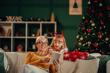 Grandmother reading christmas stories to granddaughter by christmas tree