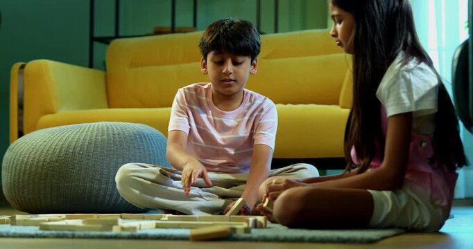 Indian Asian small sibling kids playing with wooden building blocks together, constructing toy home structure in modern living room, enjoying quality childhood activity, family bonding indoors