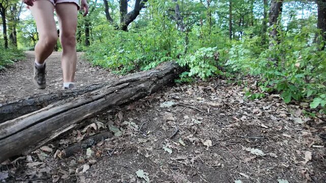 Woman's leg and hiking boot stepping over a fallen tree trunk on a dirt trail in a vibrant green forest, symbolizing active outdoor lifestyle and nature exploration. SLOW MOTION