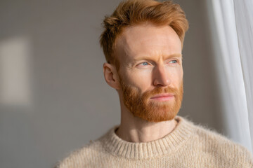 thoughtful scandinavian man with ginger beard gazes out of large window in bright serene interior