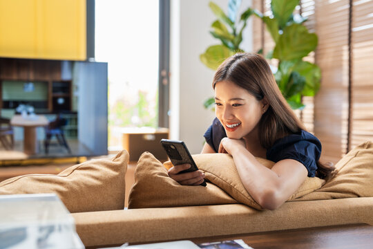 Happy pleasant woman relaxing on comfortable couch, holding smartphone in hands. Smiling young lady chatting in social networks, watching funny videos, using mobile applications at home. - Powered by Adobe